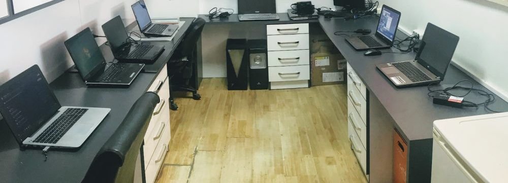 Bilgisayar Borsası atölyesindeki çalışma tezgâhı: sıra hâlinde açık duran laptoplar, duvardaki raflarda yedek parça kutuları.